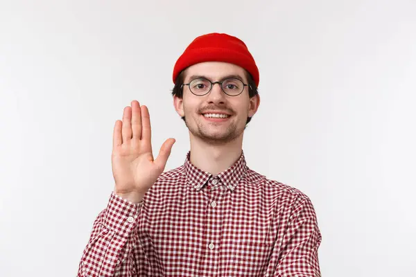 Närbild porträtt av ung söt kaukasier kille säger hej till ny lagmedlem, höja handen i hälsning gest, leende vänlig, försöker vara trevlig, bär röd mössa och glasögon, vit bakgrund — Stockfoto