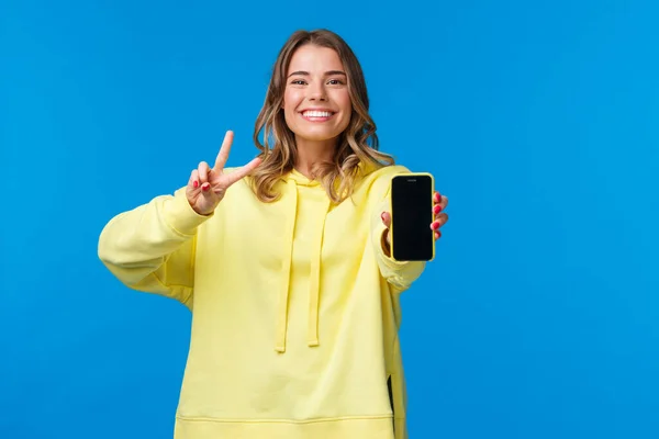 Söt leende glad blond flicka i gul luvtröja visar dig hennes online blogg eller sociala medier sida på mobiltelefon display, göra fred gest och grinar, stå blå bakgrund — Stockfoto