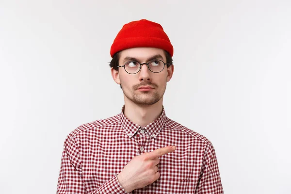 Cético bem parecido jovem hipster cara em gorro vermelho e verificar camisa, óculos, espreitando duvidoso, olhando apontando canto superior direito com expressão séria, de pé fundo branco — Fotografia de Stock