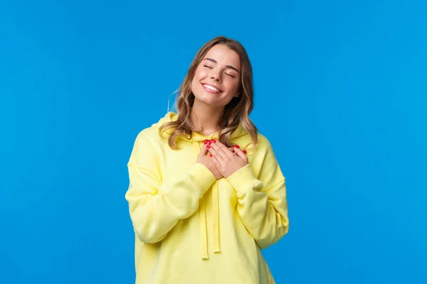 Romántica seductora novia rubia caucásica en sudadera con capucha amarilla, presionar las manos al corazón y cerrar los ojos con una sonrisa encantadora recordar hermoso recuerdo, la imagen perfecta fecha, de pie fondo azul —  Fotos de Stock