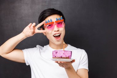 Kutlama, tatil ve doğum günü konsepti. Komik parti gözlüklü neşeli Asyalı adamın yakın plan portresi, yanan mumla doğum günü pastasını tut, barış işareti yap, dilek tut, siyah arka plan