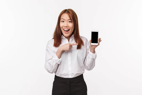 Porträt einer glücklichen eleganten jungen hübschen asiatischen Frau in weißem Hemd, stellt neue Anwendung vor oder wirbt für Firmenwebseite, Programm, zeigt mit dem Finger auf Handy, weißer Hintergrund — Stockfoto