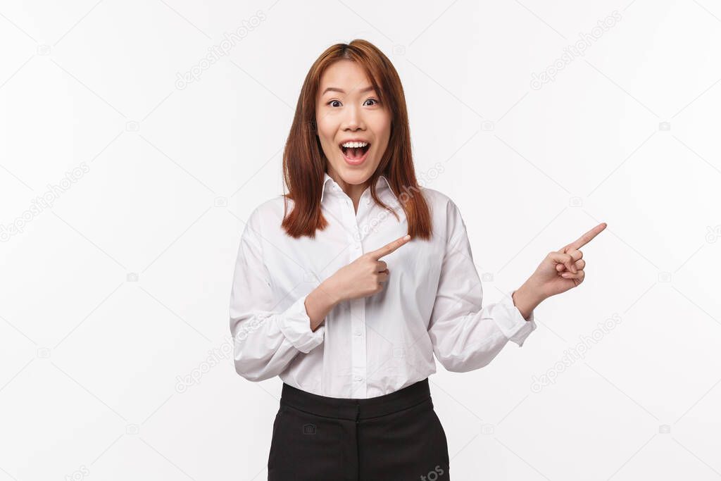 Portrait of excited wondered young asian woman in shirt and skirt, pointing fingers right smiling camera with thrilled enthusiastic expression, talking about new awesome product, white background