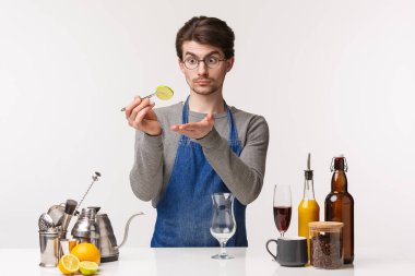 Barmen, kafe çalışanı ve barmen konsepti. Beyaz önlüklü genç beyaz erkek portresi limon dilimi tutuyor, kokteyl hazırlıyor, bardakta içki yapıyor, meyvelere bakıyor, beyaz arka plana bakıyor.