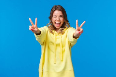 Olumlu düşün. Kawaii neşeli, arkadaş canlısı, sarı kapüşonlu, gülümseyen sarışın kadın, barış hareketleriyle ellerini uzat, dilini göster ve sırıt, heyecanlı ve neşeli hisset.