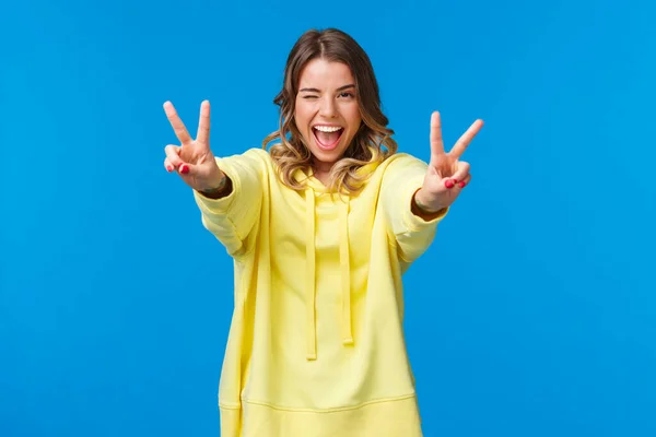 Mantém a certeza. Kawaii alegre, de aparência amigável sorrindo mulher loira com capuz amarelo, esticar as mãos para a frente com gestos de paz, mostrar língua e sorrir, sentir animado e otimista — Fotografia de Stock