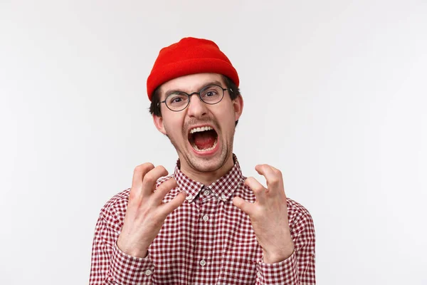 Close-up retrato de angustiado e irritado jovem hipster engraçado cara com bigode usar gorro vermelho, óculos, apertar as mãos irritado e incomodado, gritando de sentimento irritado, fundo branco — Fotografia de Stock