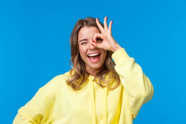 İyimser, hayalperest, kısa saçlı, sarışın bir kızın yakın plan portresi. Göze güzel bir jest yapıyor ve mutlu göz kırpıyor. Olumlu, coşkulu, mavi arka planda duruyor.