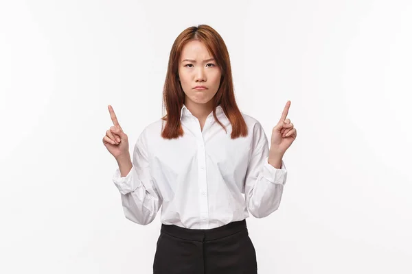 Portrait of disappointed and angry, displeased asian woman in white shirt, skirt, pointing fingers up and frowning camera upset, scolding person for bad project, awful result, white background — Stok fotoğraf