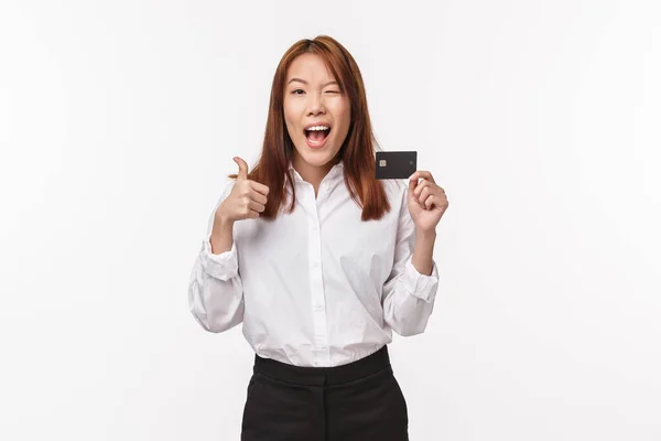 Портрет задоволеної і впевнено виглядаючої азіатки в сорочці підморгнути на камеру задоволений, показати великий палець і кредитну картку, порекомендувати банківське обслуговування, в захваті від якості, білий фон — стокове фото