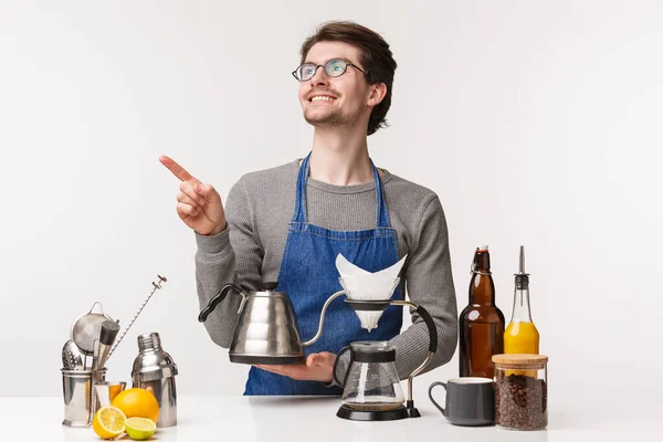 Barista, trabajador de cafetería y camarero concepto. Retrato de hombre barbudo sonriente guapo de ensueño en delantal, sosteniendo hervidor de agua y señalando esquina superior izquierda con la cara complacida . — Foto de Stock