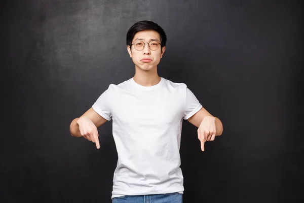 Miserabel och dyster upprörd söt asiatisk kille snyftande titta på kameran med ånger och sorg, peka finger ner på något brutet eller upprörande, känna sig orolig, svart bakgrund — Stockfoto