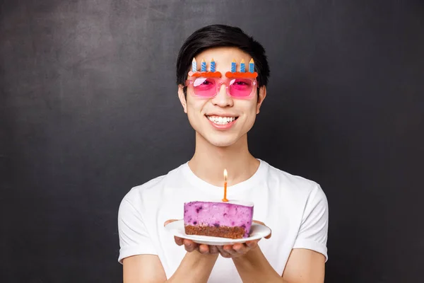 Celebration, holiday and birthday concept. Close-up portrait of excited and happy, cheerful asian man celebrating b-day in funny glasses, holding cake, blow-out candle, black background