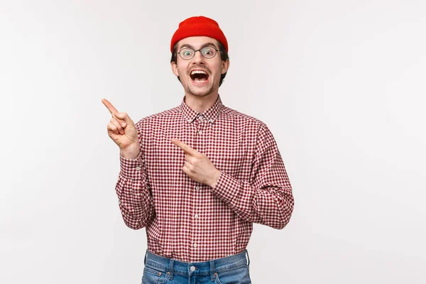 Carismático engraçado jovem em óculos e gorro vermelho introduzir algo incrível, rindo e sorrindo animado apontando dedos canto superior esquerdo, recomendo clique ou subscrever — Fotografia de Stock