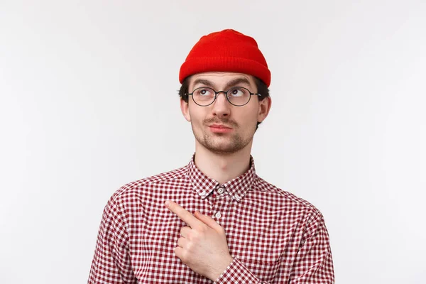 Primer plano retrato escéptico y dudoso chico barbudo guapo en gorro rojo, camisa de cuadros y gafas, sonrisa insatisfecha y vacilante apuntando esquina superior izquierda en algo malo y poco impresionante — Foto de Stock