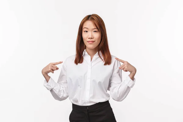 Portret odnoszącej sukcesy profesjonalnej i pewnej siebie azjatki w białej koszuli, wskazującej siebie i uśmiechniętej zadowolonej, chwalącej się własnym osiągnięciem, mówiącej o osobistych celach, stojącej na białym tle — Zdjęcie stockowe
