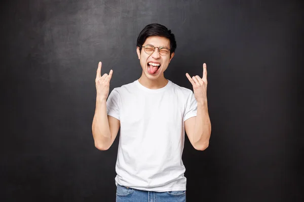 Blij opgewonden en uitgaande aziatische man in bril en t-shirt, plezier show tong sluiten ogen ontspannen en maken rock-n-roll heavy metal gebaar als het luisteren naar geweldige lied, zwarte achtergrond — Stockfoto