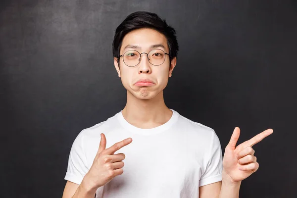 Närbild porträtt av imponerad asiatisk man i glasögon vit t-shirt, sura och ser kamera med godkännande, pekar finger rätt på riktigt cool awesome produkt, håller med dig, svart bakgrund — Stockfoto