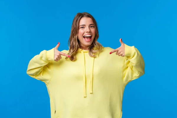 Confiante, amigável e sorridente menina loira com capuz amarelo apontando para si mesma, sugerir própria ajuda, quer voluntário ou particapte, dizer me escolher, de pé fundo azul — Fotografia de Stock