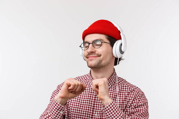 Just vibing. Happy carefree young hipster guy with moustache in glasses and red beanie dancing joyfully, listening to favorite song in headphones with pleased relaxed face, standing white background