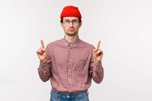 Waist-up portrait upset and disappointed whining adult man in red beanie, glasses, pointing fingers up at advertisement and showing dislike and uneasy feelings with face expression, white background
