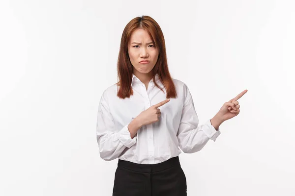 Portrait of displeased and unsatisfied asian young woman in shirt, grimacing and frowning with disgust and disappointment, pointing right at something upsetting and bothering, white background — Stock Photo, Image