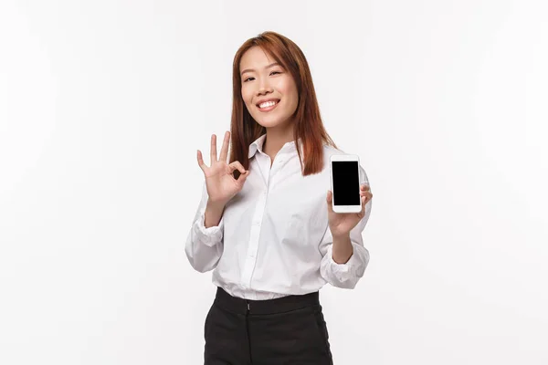 Das ist gut. Zufrieden gut aussehende asiatische Frau zeigt Handy-Display und machen okay Geste, bewerten ausgezeichnete App, stolz auf die Aufnahme von coolen Foto, stehen weißen Hintergrund — Stockfoto