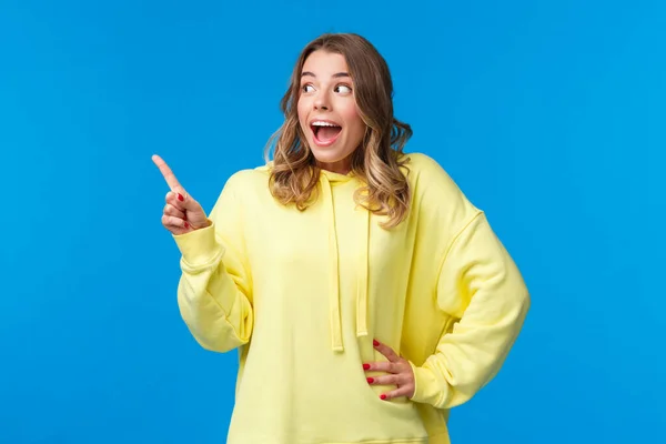 Garota loira feliz sem palavras animado reagir a notícias impressionantes, apontando e olhando para o espaço de cópia em branco esquerdo como se estivesse vendo algo impressionante e legal, de pé fundo azul — Fotografia de Stock