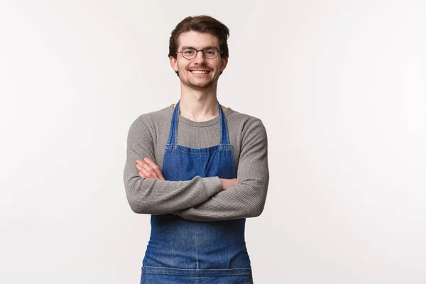 Cintura-up retrato de alegre amigável jovem barista no avental, cruzar as mãos sobre o peito como um profissional, sorrindo feliz, cumprimentando os clientes como estando na cafetaria ou bar balcão — Fotografia de Stock