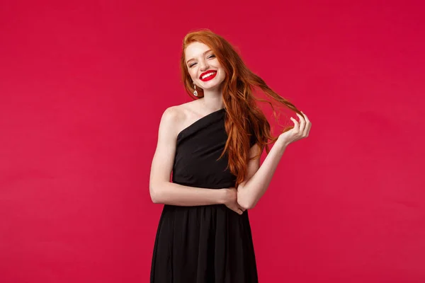 Elegancia, moda y concepto de mujer. Hermosa mujer joven pelirroja guapa en vestido elegante negro, coqueta sonriendo con emoción complacida, hilo de pelo ondulado coqueta, fondo rojo — Foto de Stock