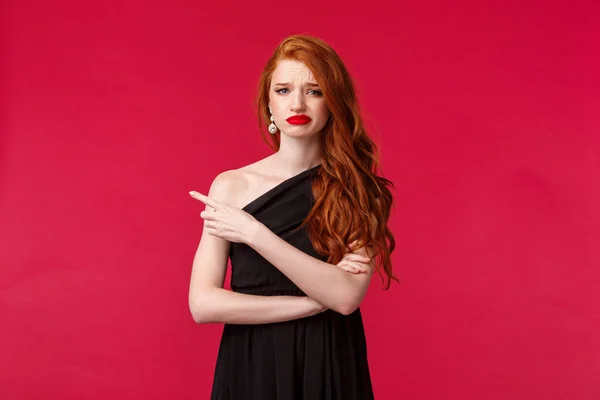 Portret van sceptische, kieskeurige elegante roodharige vriendin in stijlvolle zwarte jurk, grimmig teleurgesteld, wijzende vinger links naar iets slechts, afkeer en oneens, staan rode achtergrond — Stockfoto