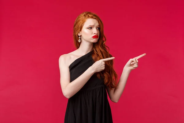 Celebração, eventos, conceito de moda. Retrato de triste sombrio jovem mulher ruiva elegante em belo vestido preto olhar decepcionado, franzindo a testa e sorridente chateado no lado direito promo, fundo vermelho — Fotografia de Stock