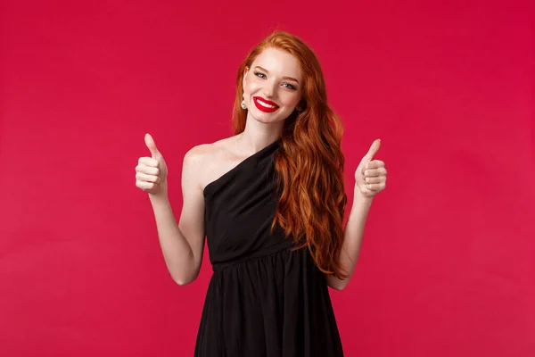 Conceito de moda, luxo e beleza. Retrato de encantador apoiante jovem ruiva em preto elegante vestido, maquiagem, sorrindo agradou mostrar polegares-up em aprovação ou como, fundo vermelho — Fotografia de Stock