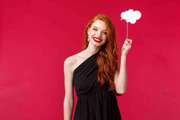 Retrato de encantador elegante jovem ruiva mulher em vestido preto, segurando comentário nuvem vara sobre a cabeça e sorrindo, à procura de inspiração, ter ideia, stand fundo vermelho — Fotografia de Stock