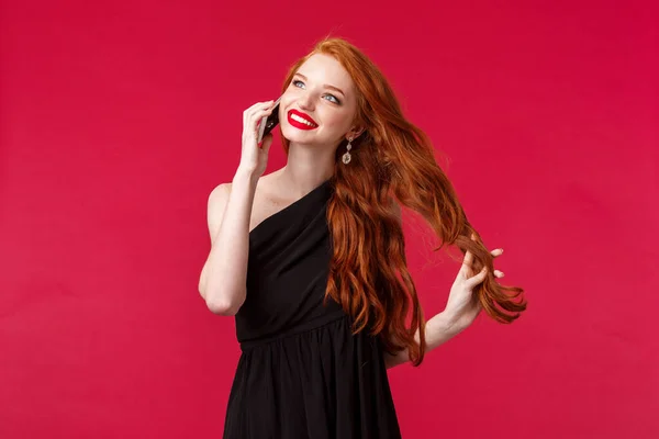 Retrato de hermosa, elegante joven pelirroja con el pelo largo y rizado de jengibre, tocando hebra despreocupada hablando por teléfono, recibir elogios durante la conversación, sonriente mirada de ensueño en vestido negro — Foto de Stock