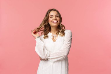 Güzel ve kendine güvenen genç, sarışın, hoş, şık, beyaz elbiseli, saçlarına dokunan ve gülümseyen, romantik randevu için giyinen, kadınlar ve güzellik konsepti, pembe arka plan