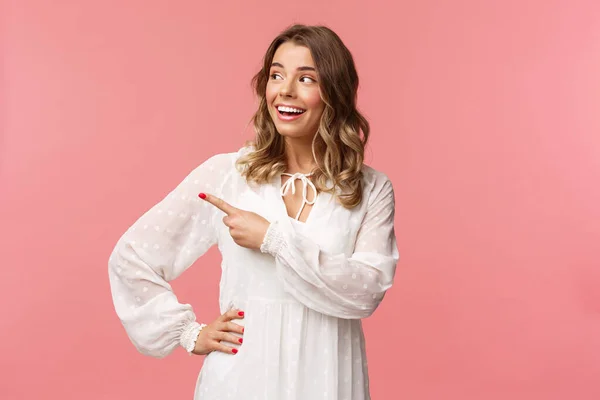 Curiosa mujer rubia hermosa optimista disfrutando de la sensación de verano alegre, apuntando mirando a la izquierda con expresión interesada, entusiasta, usar lindo vestido blanco, de pie fondo rosa — Foto de Stock