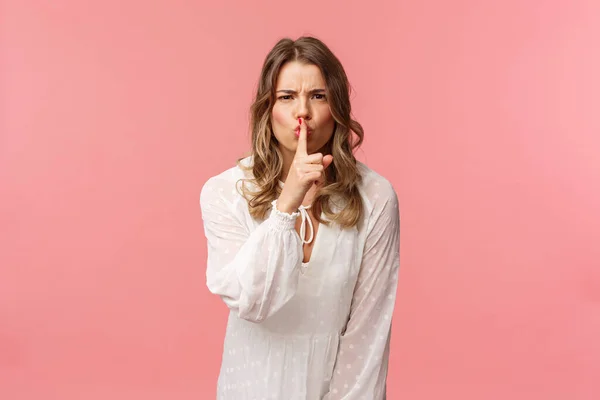 Beauty, fashion and women concept. Angry serious-looking blond girl in white dress, shushing at someone making noise, press finger to lips scolding bad behaviour, say be silent, pink background — Stock Photo, Image