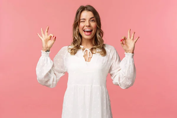 Beauty, fashion and women concept. Portrait of carefree unbothered blond feminine woman in white dress, guarantee quality, wink say no problem, smiling pleased, rate good product, pink background — Stock Photo, Image