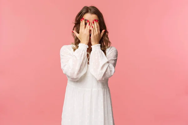 Portret van dom schattig meisje met blond kort haar, draag witte jurk, verbergen gezicht achter de handen, beloven om te wachten op signaal, maar gluren door vingers, kan niet weerstaan verleiding, staan roze achtergrond — Stockfoto