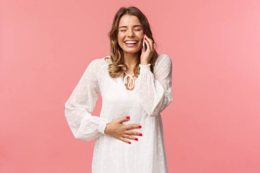 Neşeli, mutlu, beyaz elbiseli genç bir kızın portresi, eğleniyor, arkadaşıyla konuşuyor, cep telefonunu arıyor, gülüyor ve karnına dokunuyor, gülümsüyor, akıllı telefonu kulağının yanında tutuyor.