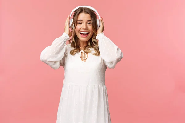 Retrato de animado, menina de boa aparência feliz em vestido macio branco, usando fones de ouvido e sorrindo espantado como olhar para a câmera, fascinado com boa qualidade de som, fundo rosa — Fotografia de Stock