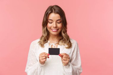 Beyaz elbiseli, heyecanlı ve neşeli sarışın kızın yakın plan portresi, elinde kredi kartı ve gülümseyen heyecanla, bir şeyler satın alma arzusuna karşı koyamaz, online alışveriş için para israfı, pembe arka plan