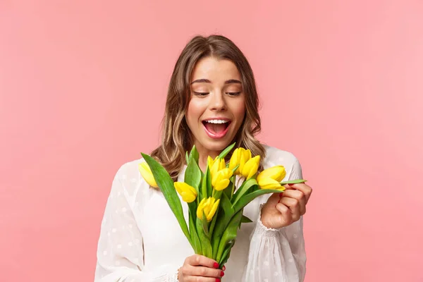 Primavera, felicità e concetto di festa. Ritratto di ragazza felice sorpresa e contenta guardando allegramente il mazzo di fiori, sorridente divertito, ricevere tulipani gialli, stare sullo sfondo rosa — Foto Stock