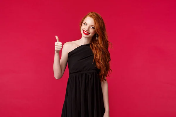 Elégance, mode et concept de femme. Portrait de charmante jeune femme rousse élégante satisfaite en robe élégante noire sur fond rouge, montrer pouces vers le haut dans l'approbation ou comme signe — Photo