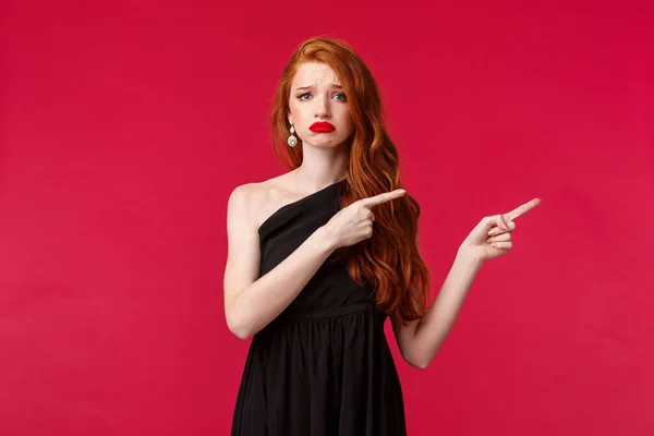 Celebración, eventos, concepto de moda. Retrato de mujer pelirroja bonita incómoda y triste en vestido negro, mueca y frunciendo el ceño angustiado, sentir dolor o arrepentimiento, señalando los dedos a la derecha, fondo rojo — Foto de Stock