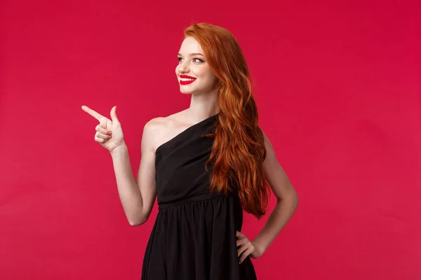 Retrato de sensual y femenina joven pelirroja en elegante vestido negro, asistir a la fiesta, espectáculo de alfombras rojas, señalando con el dedo a la izquierda y buscando satisfecho con la sonrisa feliz, radiante sonrisa — Foto de Stock