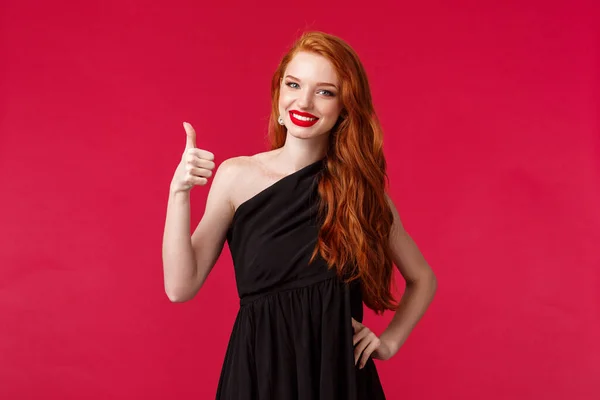 Conceito de moda, luxo e beleza. Retrato de menina elegante ruiva satisfeito, assertiva em vestido preto, mostrar polegares para cima na aprovação ou gesto semelhante, sorrindo aprovadamente, fundo vermelho — Fotografia de Stock