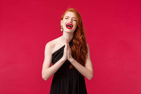 Retrato de la joven pelirroja desesperada sombría en vestido de noche negro, lloriqueando y llorando su corazón como suplicando perdón, ojos cerrados sollozando, rogando ayuda, fondo rojo — Foto de Stock