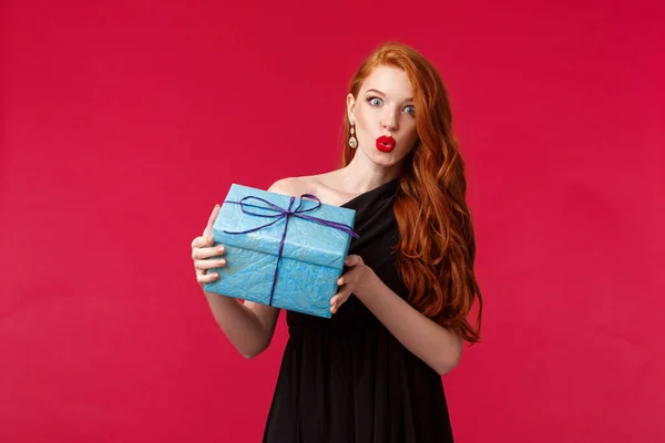 I wonder whats inside. Curious stunning young redhead woman in elegant black dress, shaking box with gift, folding lips amazed and intrigued look camera, receive present, red background